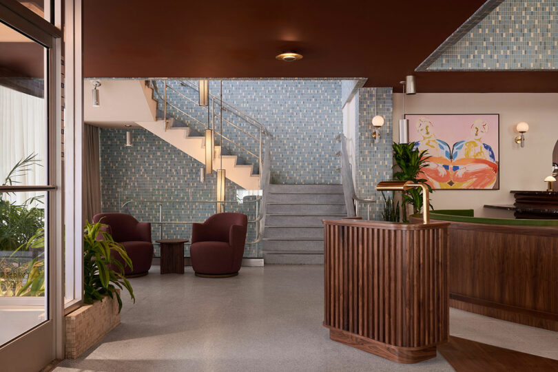 A modern Municipal Grand lobby features a wooden reception desk, two maroon chairs, indoor plants, a staircase with a glass railing, and a colorful abstract painting on a blue-tiled wall.