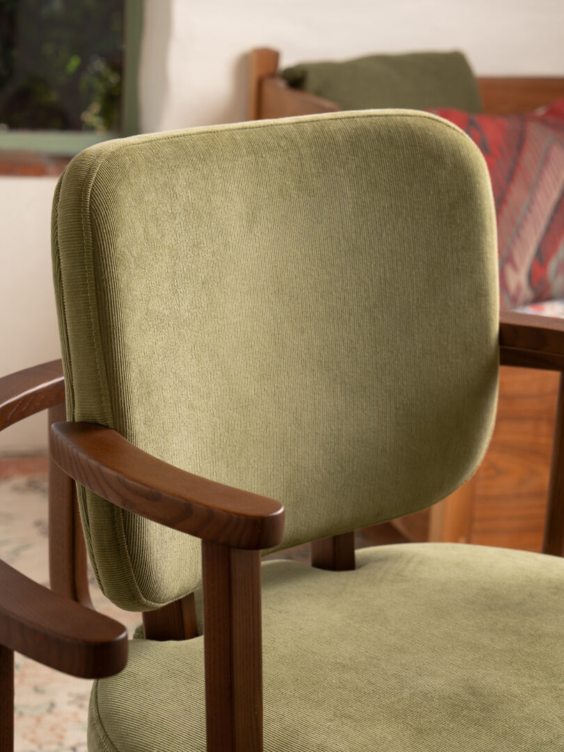 Green upholstered chair with wooden arms and legs positioned in a room with a bed and bedside table in the background.