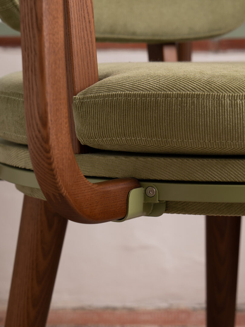 Close-up of a chair with a green corduroy cushion, wooden legs and armrest, and a green metal support bracket underneath the seat.