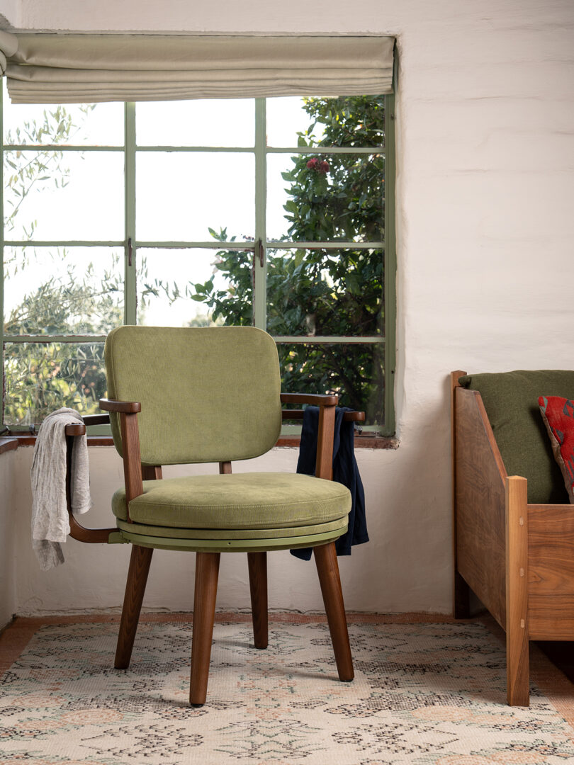 A green cushioned wooden chair sits on a patterned rug near a window with greenery outside; a white scarf and blue garment hang on the chair's arms.