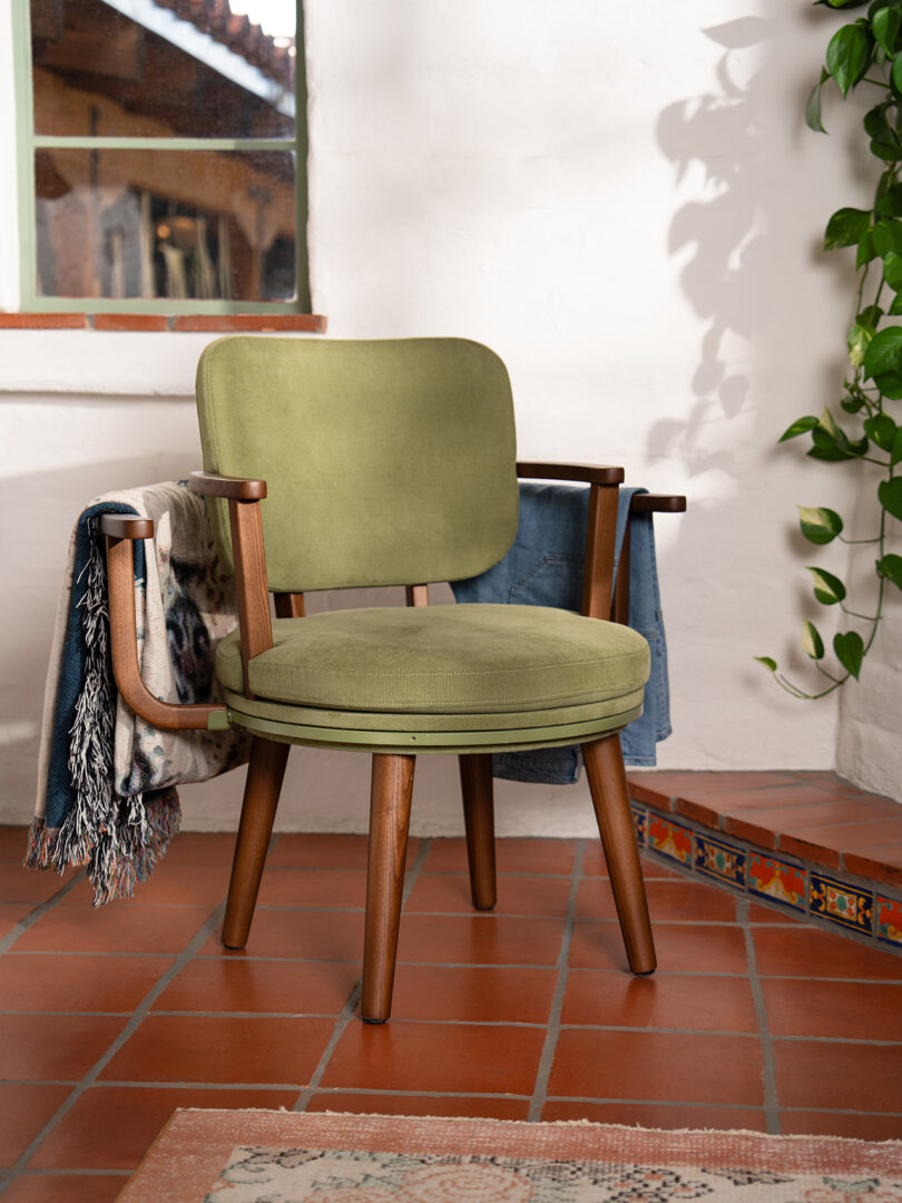 A green cushioned wooden chair sits on a tiled floor near a wall, with blankets draped over its arms and a leafy plant hanging nearby.