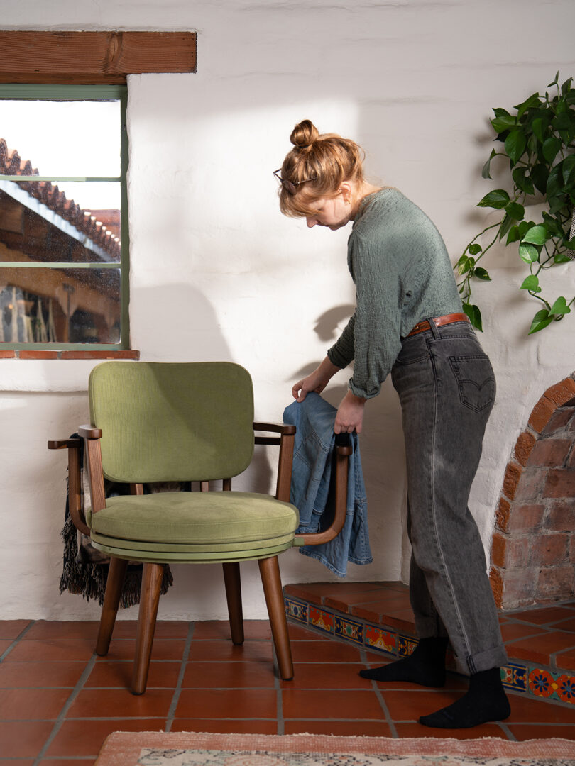 A woman stands next to a green chair in a sunlit room, draping a denim jacket over the chair’s arm.