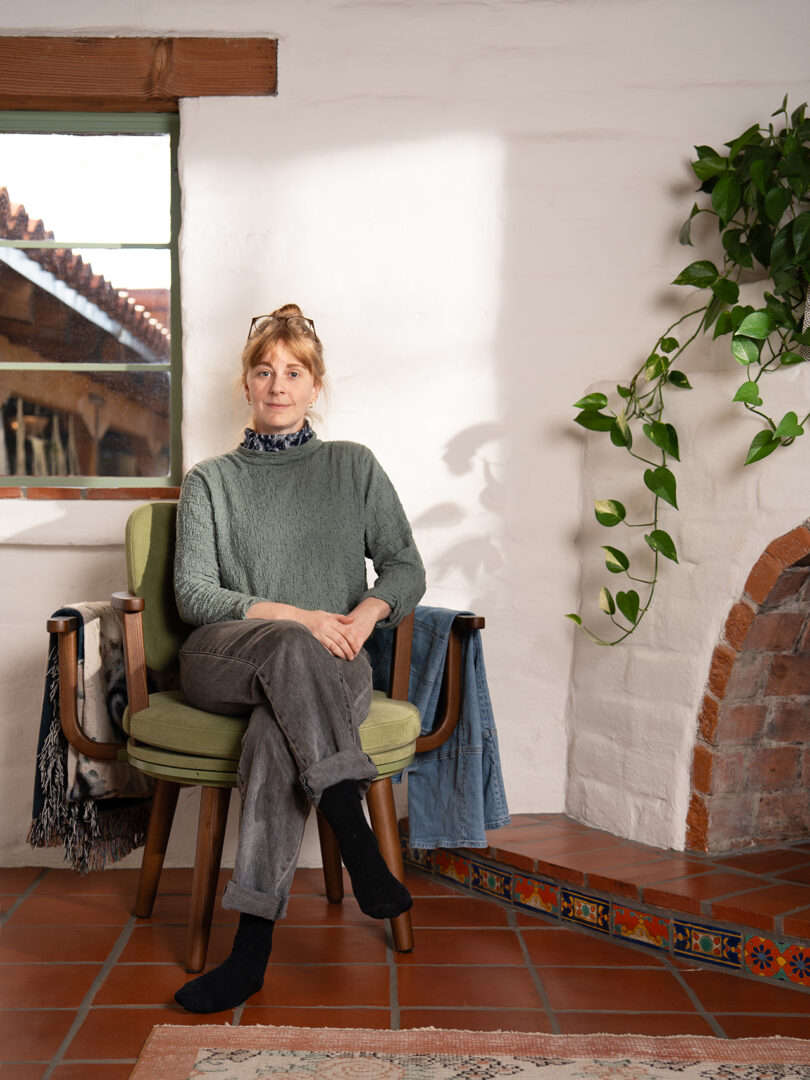 A woman sits on a green chair in a cozy room with terracotta tile floor, a brick fireplace, and a hanging plant. She wears a green sweater, gray pants, and black socks.