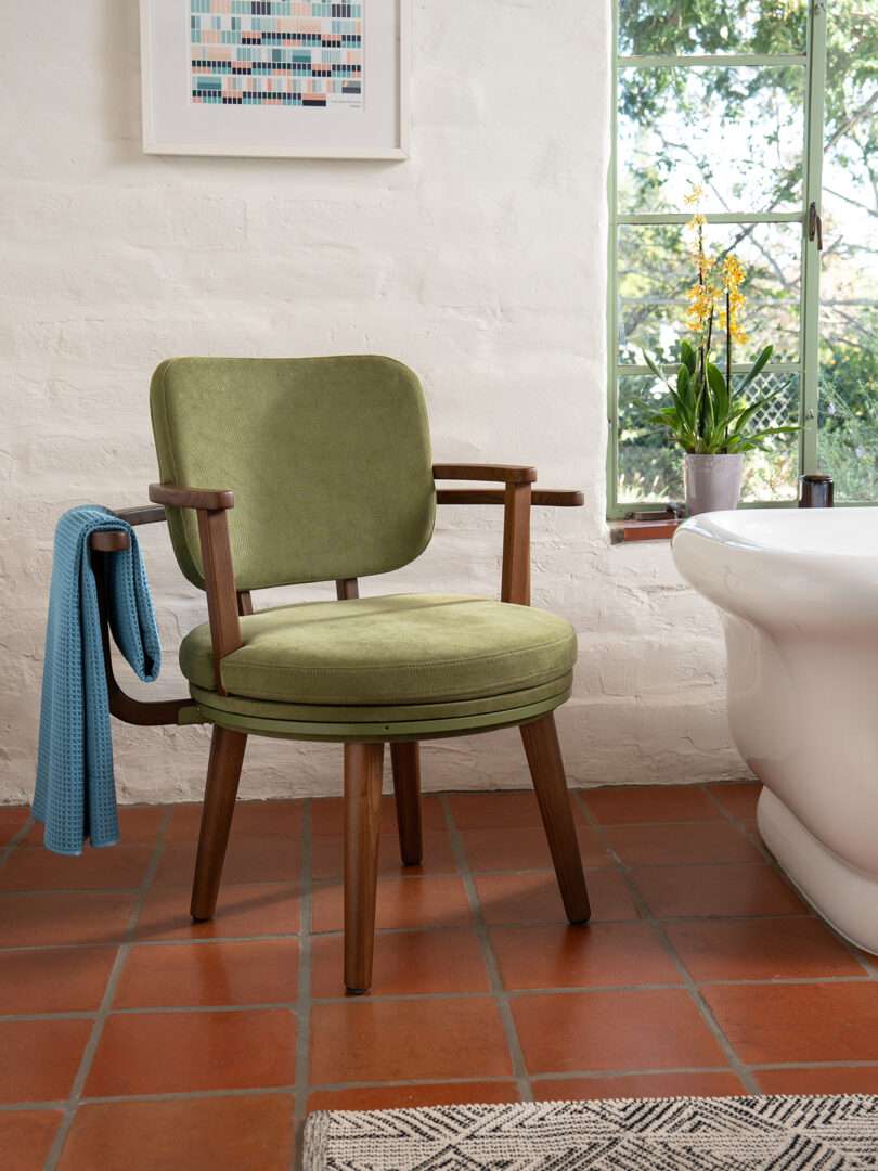 A green cushioned wooden chair is placed on a terracotta tile floor next to a bathtub; a blue towel hangs over the chair’s arm. A window and flower pot are visible in the background.