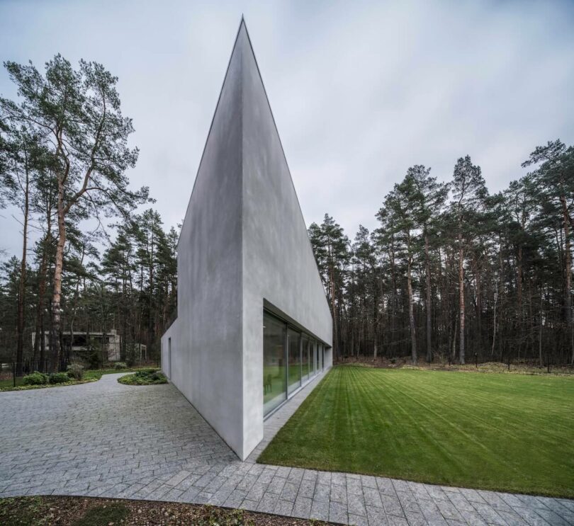 A modern, angular concrete building by KWK Promes with large windows sits on a manicured lawn next to a paved walkway, surrounded by tall pine trees.