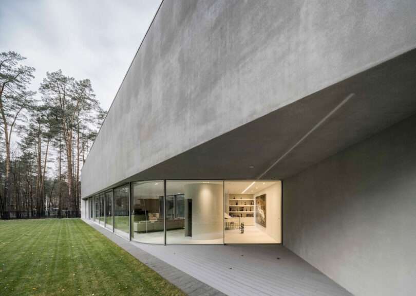Modern minimalist building by KWK Promes with large glass windows and concrete exterior, adjacent to a neatly mowed lawn and bordered by a forested area.