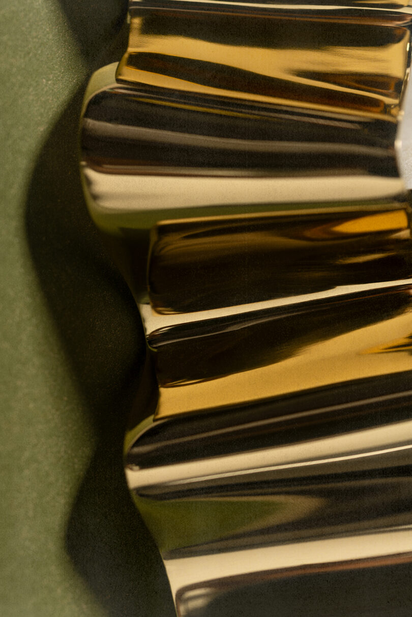 Close-up of a shiny, gold metallic gear with highlighted ridges, styled in the manner of Tali Roth, positioned against a matte green background.