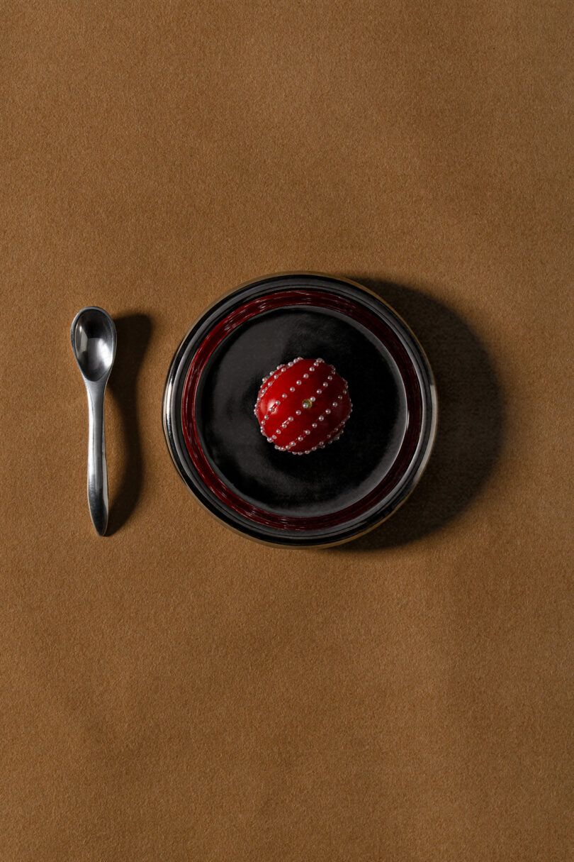 A small dessert with a shiny red dome and pearl-like decorations sits in a glass bowl filled with dark sauce, evoking Tali Roth's chic aesthetic, next to a silver spoon on a brown surface.