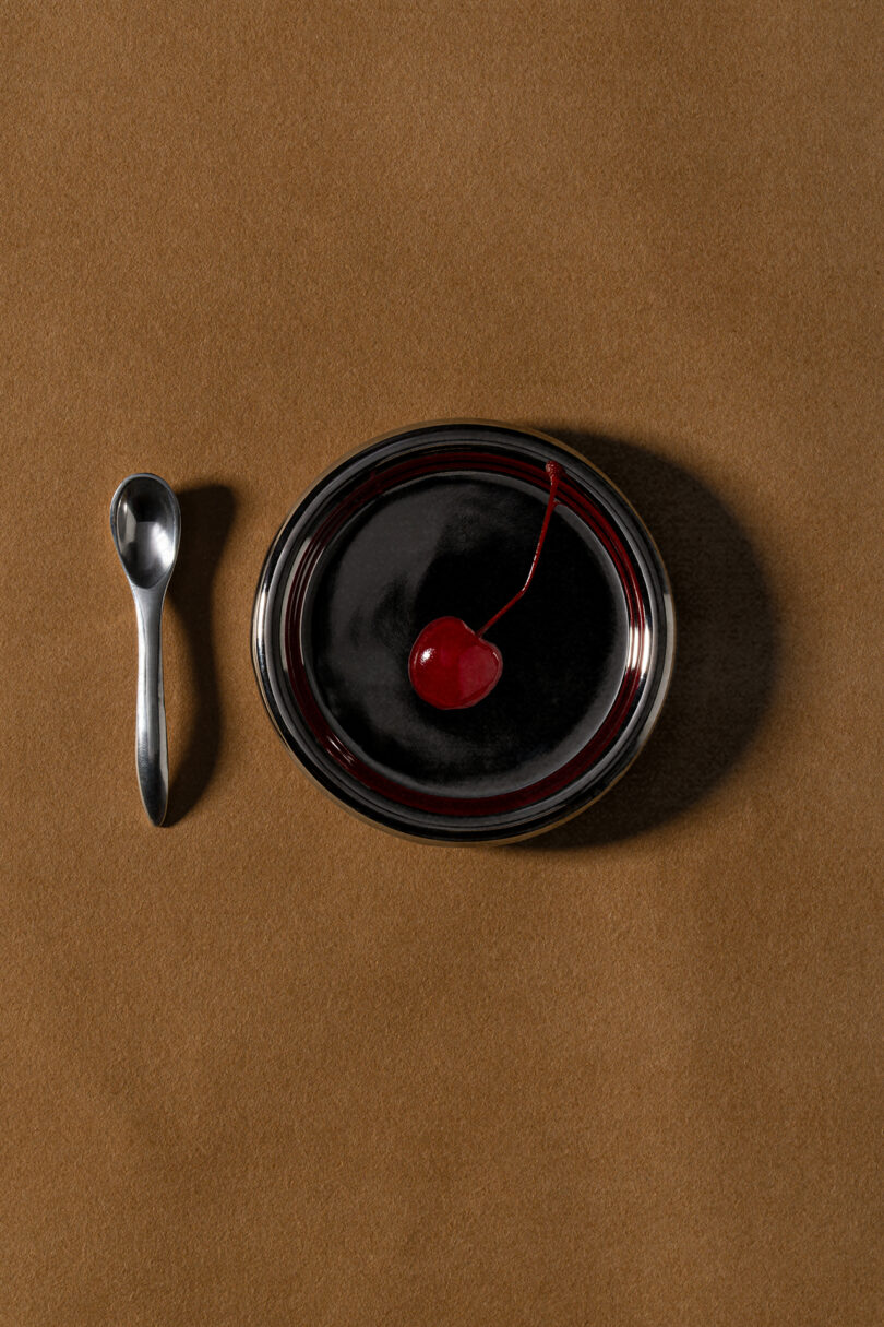 A small metal spoon beside a round dish filled with glossy dark dessert, topped with a single red cherry, rests on a brown surface in true Tali Roth style.