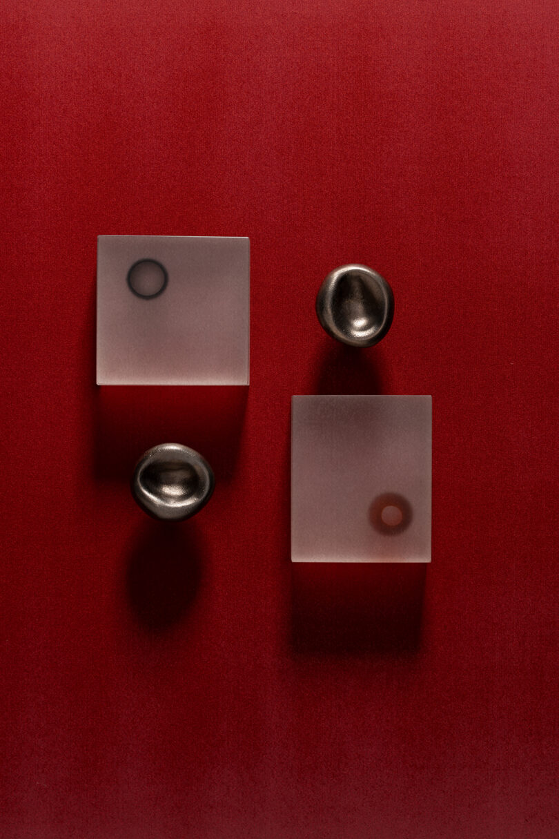 Two frosted square blocks and two small metallic bowls are arranged on a red background, with circular shadows and reflections visible on the blocks, evoking a minimalist style reminiscent of Tali Roth.
