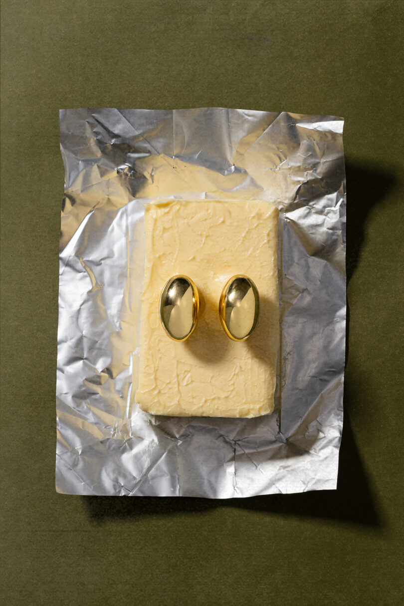 A block of butter on crumpled foil with two gold oval objects placed upright on top, styled in the distinct aesthetic of Tali Roth, all set against an olive green background.