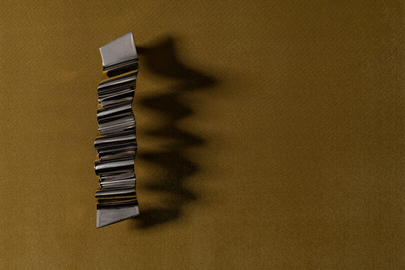 A vertically placed, folded metallic strip casts a zigzag shadow on a textured brown surface, evoking the refined minimalism often seen in Tali Roth’s interior designs.