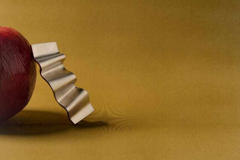 A wavy metal blade is partially inserted into a red apple on a plain brown background, reminiscent of the minimalist style favored by Tali Roth.