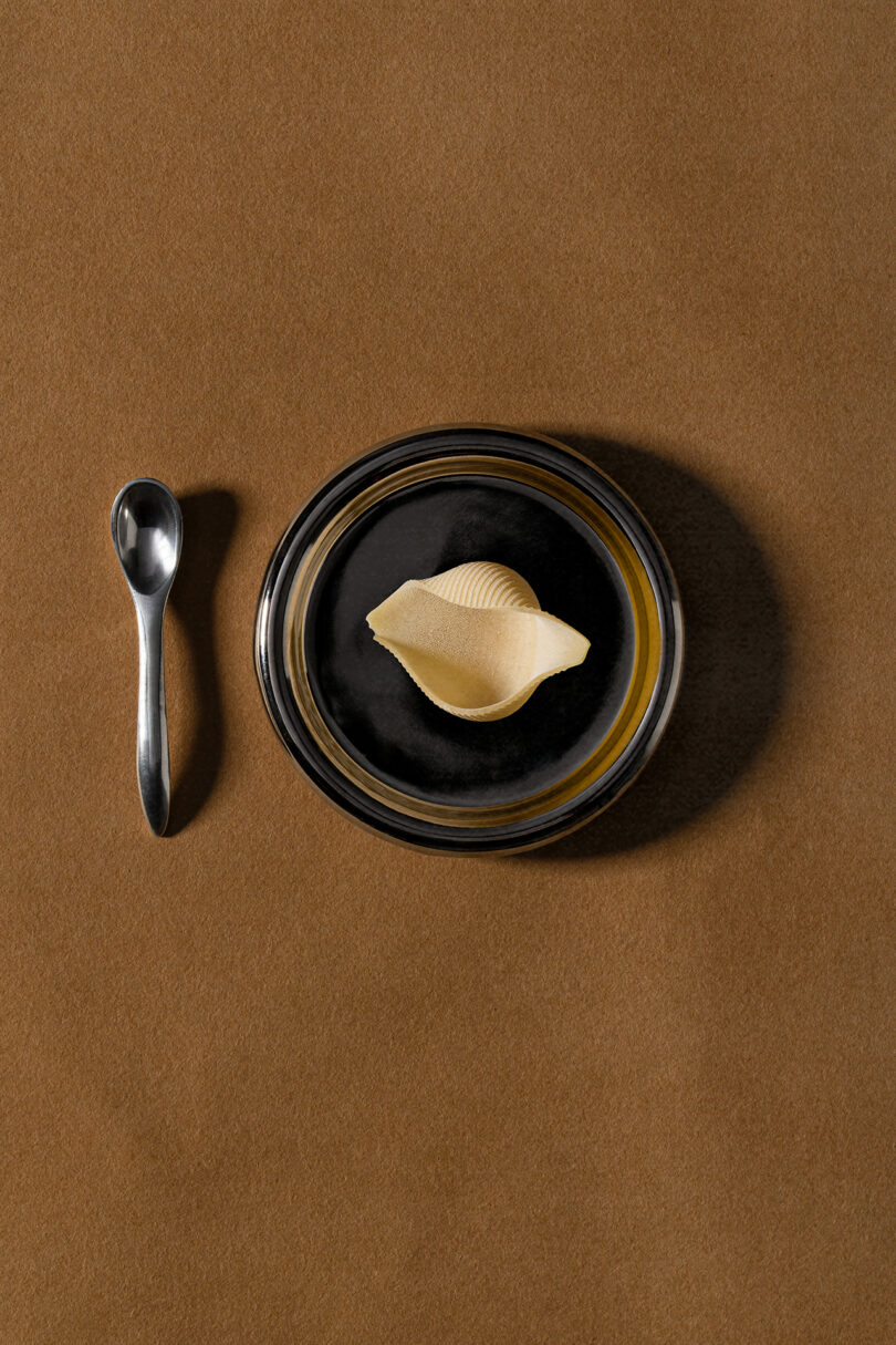 A single uncooked conchiglie pasta sits in a black bowl next to a metal spoon on a brown surface, styled with the minimalist elegance of Tali Roth.