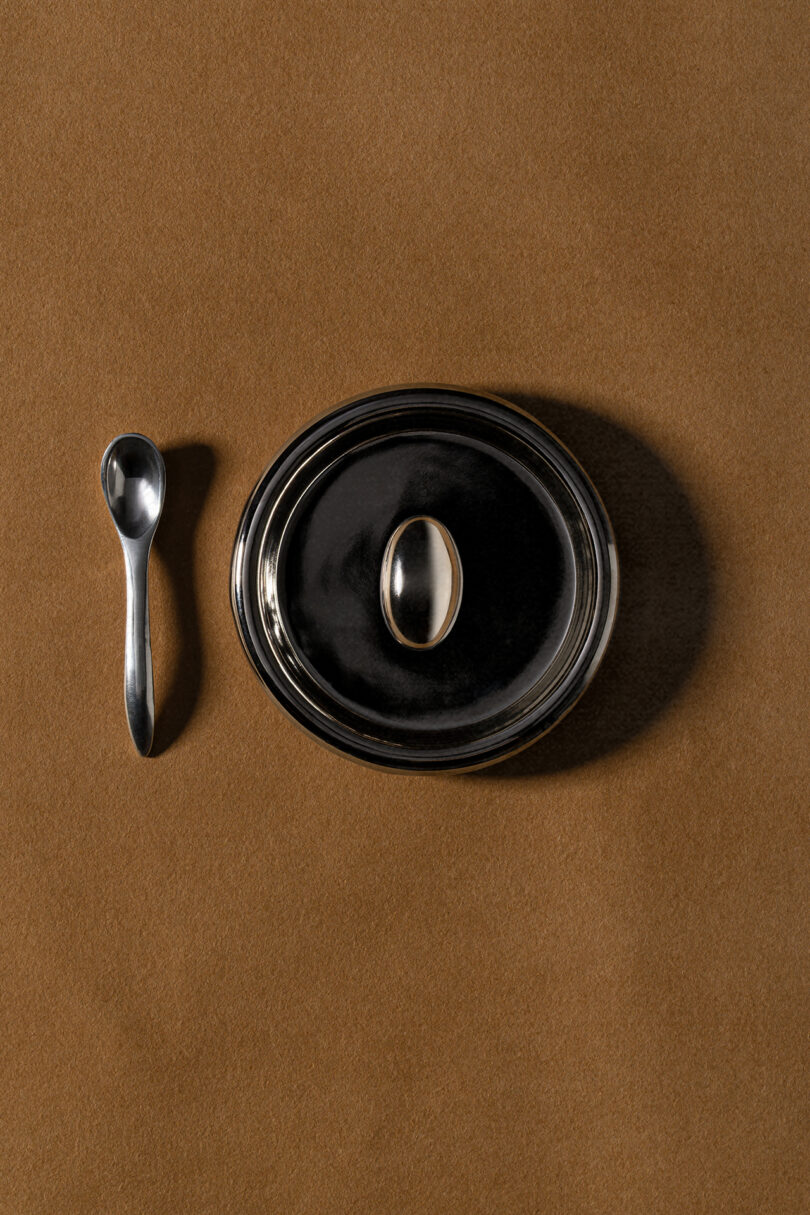 A silver spoon is placed beside a round, black-lidded dish on a brown surface, evoking the refined minimalism often seen in Tali Roth interiors.