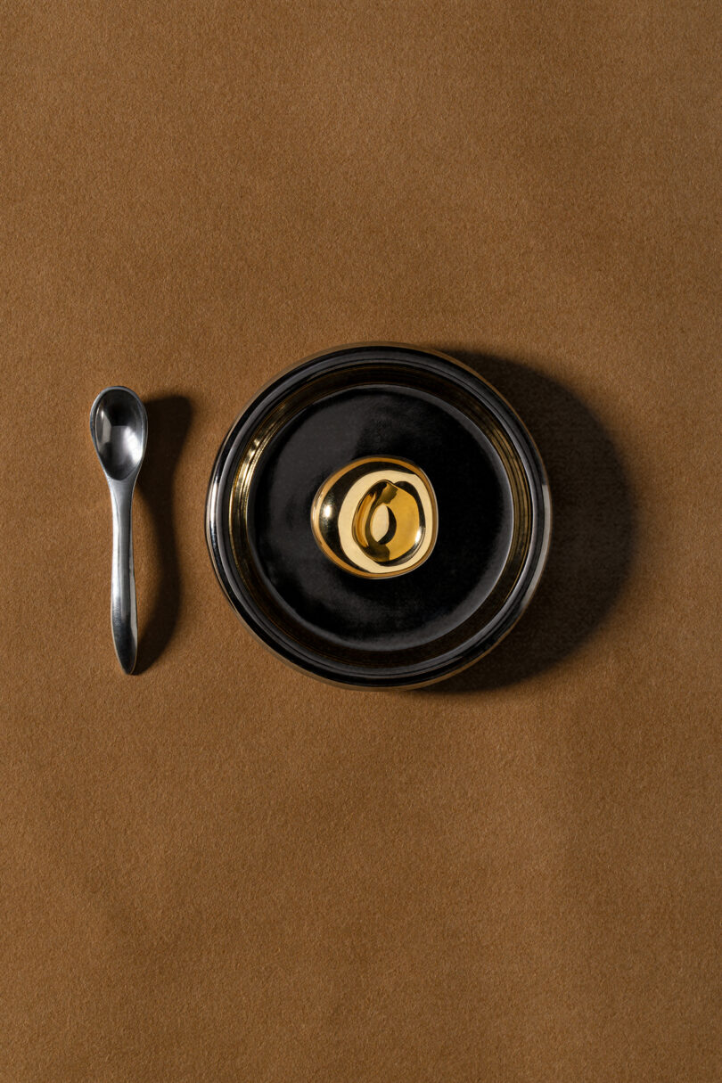 A small metal spoon and a black lidded bowl with a gold handle, styled by Tali Roth, are placed on a brown surface.