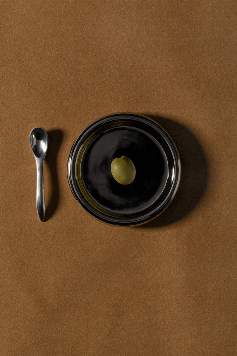 A single green grape on a black plate, placed next to a small metal spoon, rests on a brown surface in a composition reminiscent of Tali Roth’s refined aesthetic.