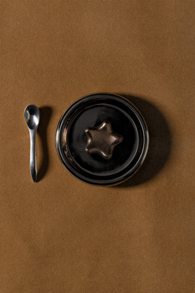 A small metal spoon beside a black bowl with a star-shaped object on top, styled by Tali Roth, placed on a brown surface.