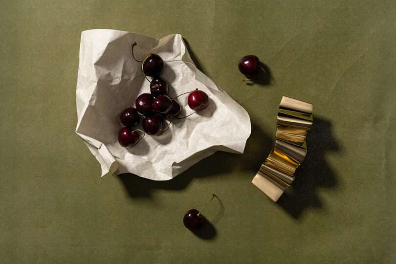 A cluster of dark cherries on crumpled parchment paper sits beside a small, gold metallic object on a green textured surface, evoking the sophisticated aesthetic often seen in Tali Roth’s curated interiors.