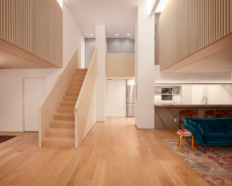 Modern open-concept interior with light wood floors, staircase, white walls, a kitchen with marble countertop, and a blue velvet sofa on a patterned rug.