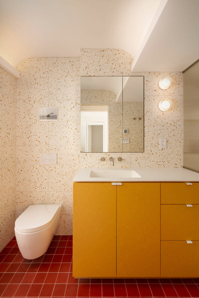 A modern bathroom with a yellow vanity, terrazzo wall tiles, a wall-mounted toilet, red floor tiles, a large mirror, and two round wall lights.