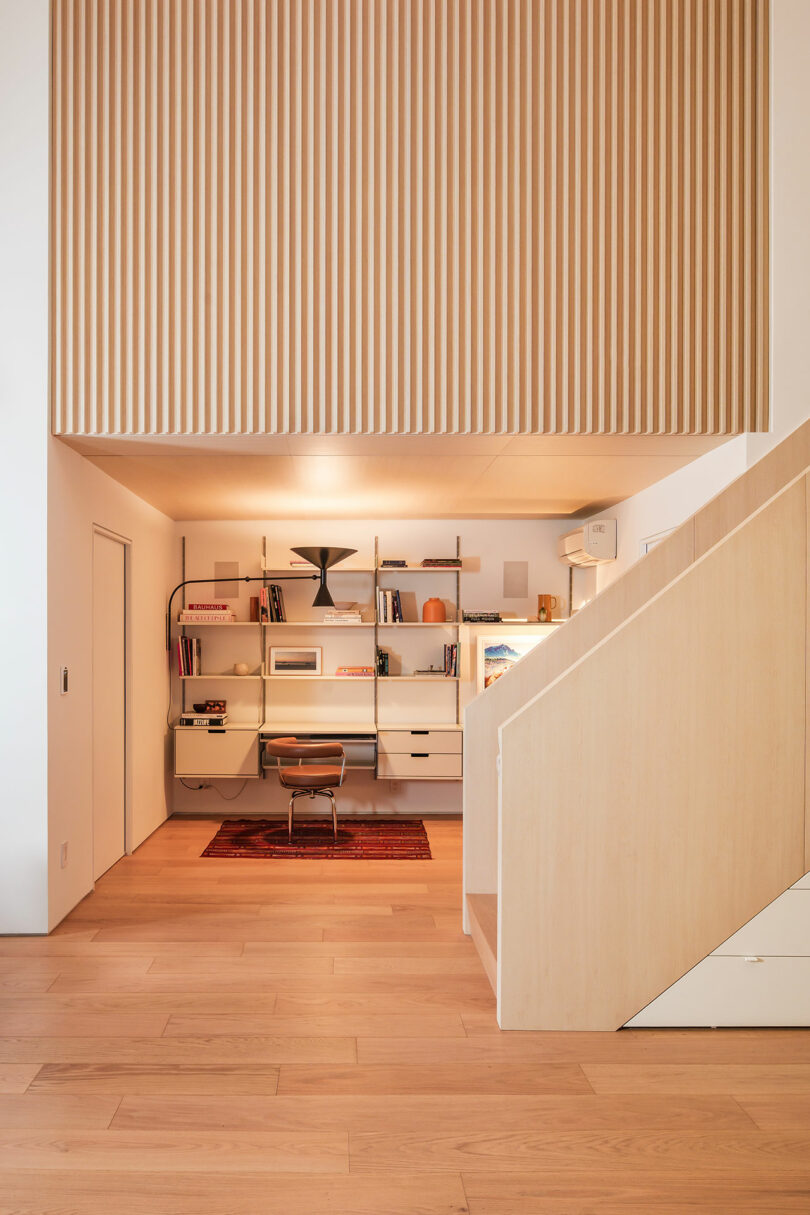 A modern home office with built-in shelves, a desk, books, decor items, and a chair beneath a wooden slatted loft area, next to a light-colored staircase.