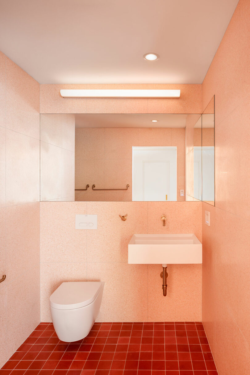 Minimalist bathroom with pink tiled walls, red tiled floor, wall-mounted toilet, white sink, brass fixtures, large mirror, and overhead light.