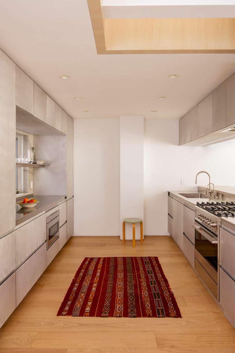 Modern galley kitchen with light wood floors, gray cabinetry, stainless steel appliances, and a red patterned rug in the center. A small stool sits against the far wall.