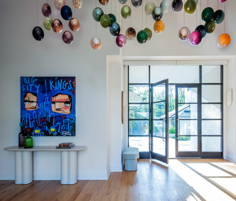 A modern entryway with glass doors, colorful hanging glass orbs, wood flooring, and a blue abstract painting above a white console table.