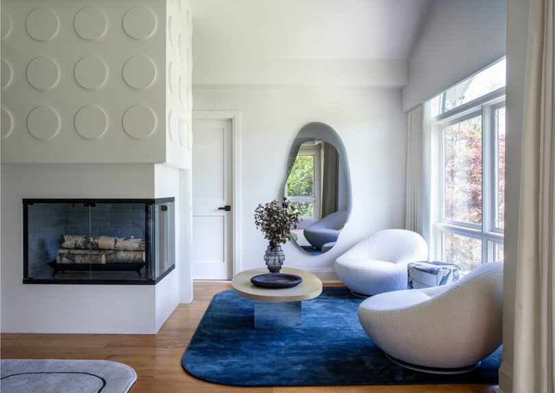 Modern living room with white sculptural chairs, oval mirror, round coffee table on a blue rug, large windows, and a corner fireplace with circular wall accents.