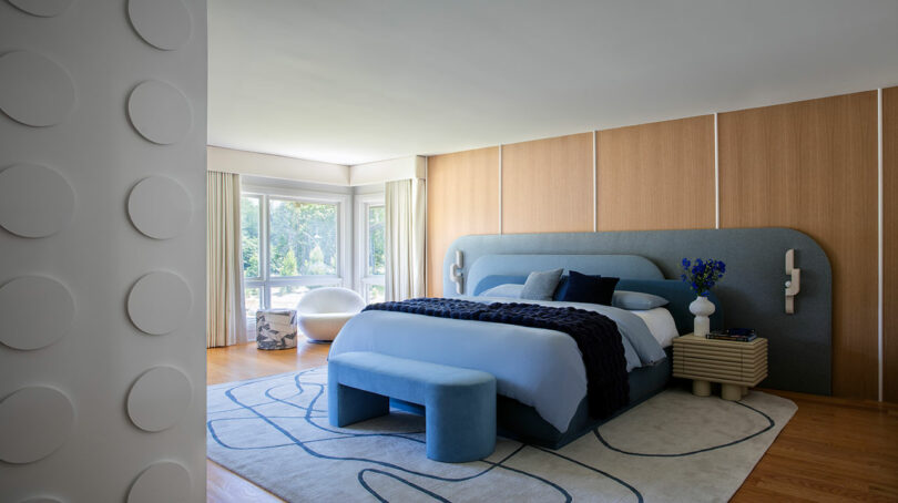 Modern bedroom with wood-paneled walls, large bed with blue accents, geometric rug, curved window area, and contemporary decor.