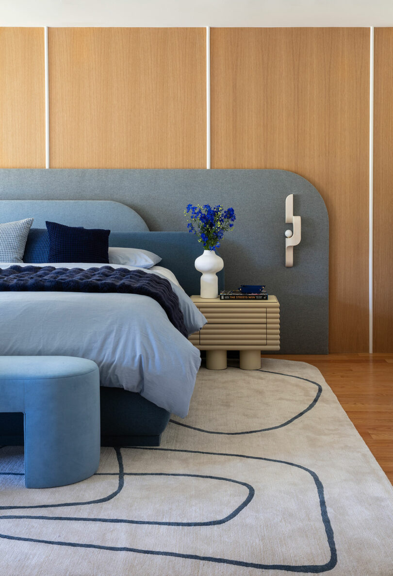 Modern bedroom with a blue upholstered bed, geometric nightstand, white vase with blue flowers, and abstract rug on a wooden floor, against a wood-paneled accent wall.