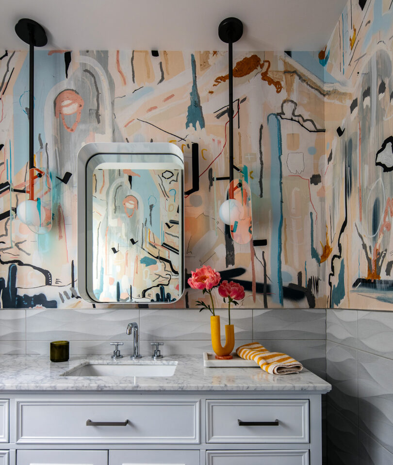 Bathroom with a marble countertop, white cabinetry, modern mirror, pendant lights, abstract wall art, and a vase with pink flowers on the counter.