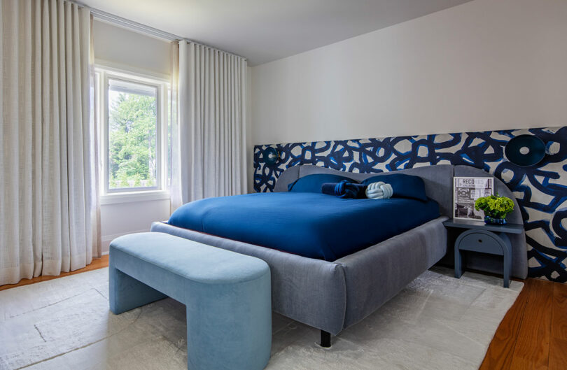 A modern bedroom with a blue and gray color scheme, featuring a bed with blue bedding, a matching bench, abstract wall art, side tables, and a large window with white curtains.