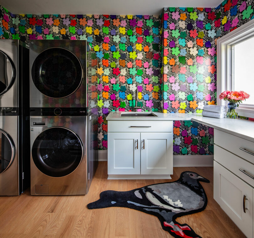 Modern laundry room with stacked washer and dryer, colorful patterned wallpaper, white cabinets, a window, folded towels, flowers, and a pigeon-shaped rug on a wood floor.