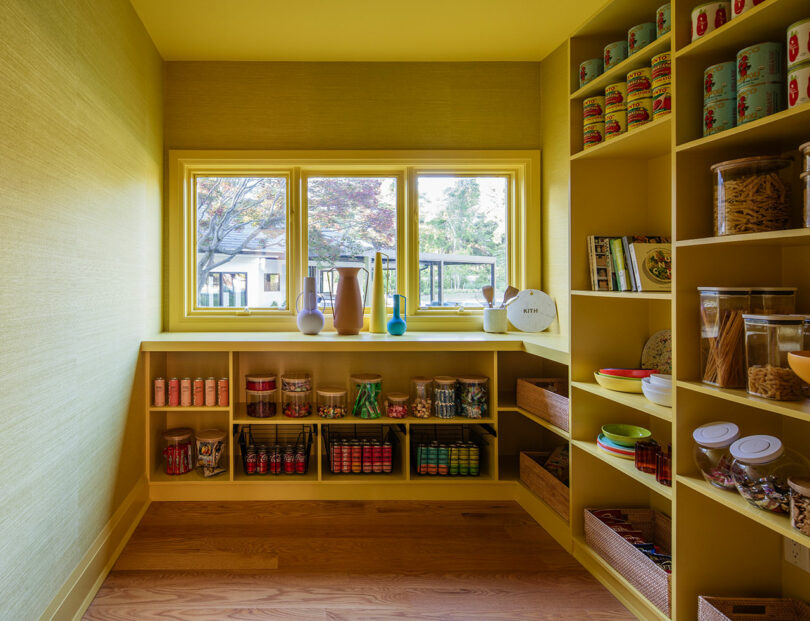 Yellow pantry with open shelves neatly stocked with jars, cans, and baskets; three windows provide natural light and a view outside.