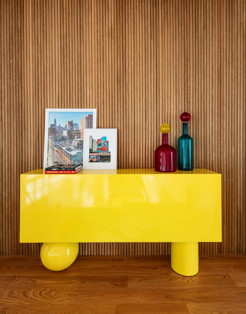 A yellow modern bench against a wooden slat wall, displaying two framed photos and three colorful glass vases on a wooden floor.