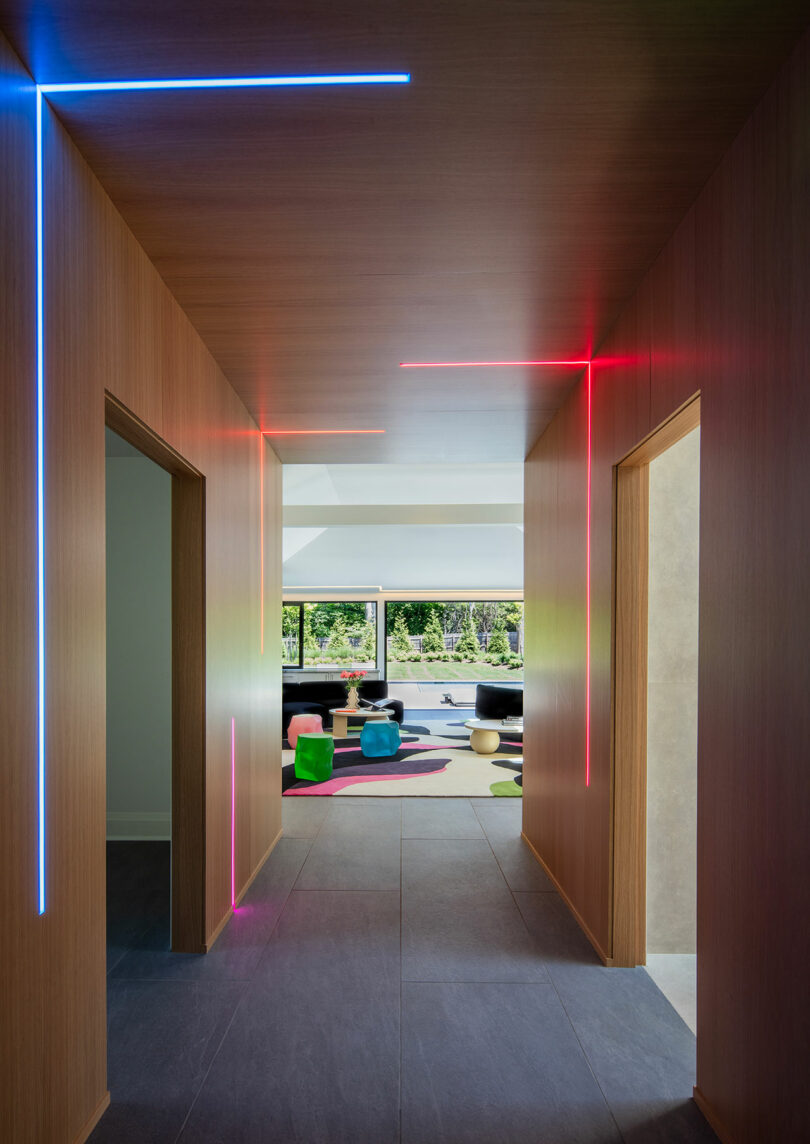 A modern hallway with wooden walls, featuring vertical and horizontal neon light strips in blue, pink, and red, leading to a bright living area with colorful furniture and large windows.