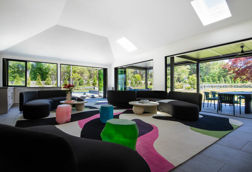 Modern living room with black sofas, colorful geometric rug, small stools, large windows, and view of outdoor patio and greenery.