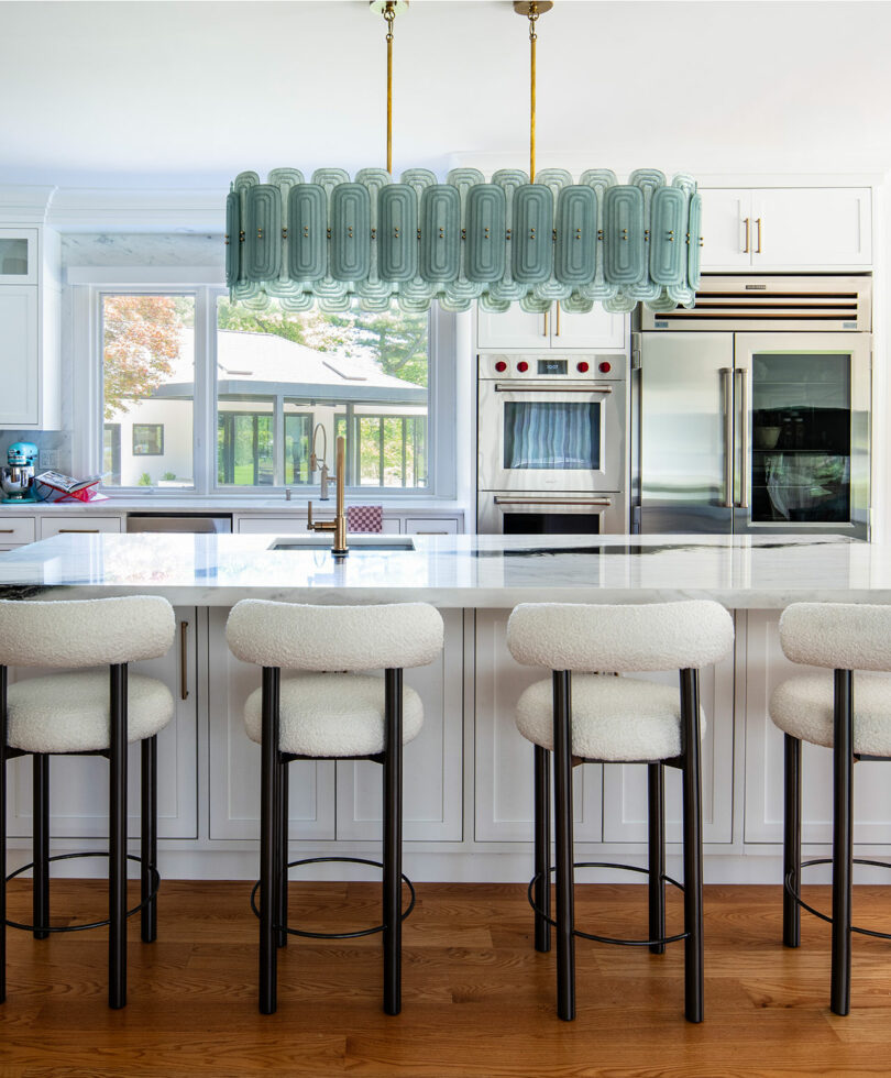 Modern kitchen with a large white island, five upholstered bar stools, brass fixtures, stainless steel appliances, and a decorative green pendant light fixture.