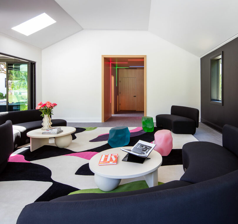 Modern living room with black sofas, colorful abstract rug, round white coffee tables, geometric stools, and bright accent decor under a vaulted ceiling with a skylight.