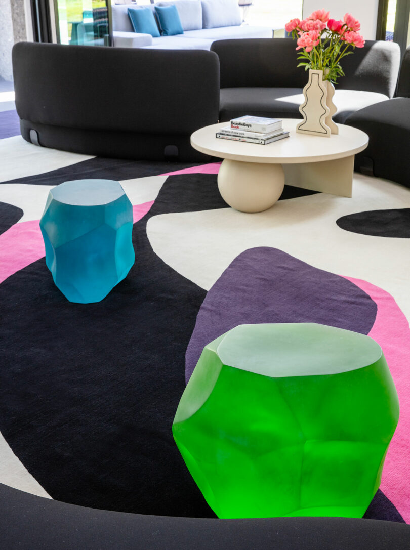 Modern living room with a curvy black sofa, geometric multicolor rug, blue and green sculptural stools, a round coffee table with books, and a vase of pink flowers.