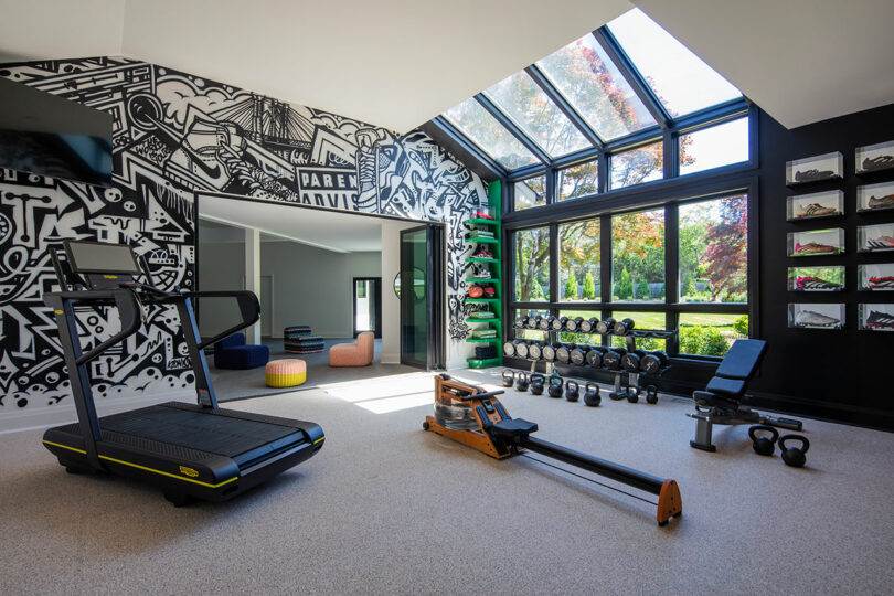Home gym with treadmill, rowing machine, weights, bench, kettlebells, wall art, and large windows overlooking a garden. Several shelves hold gym equipment and green yoga mats are stacked in a corner.