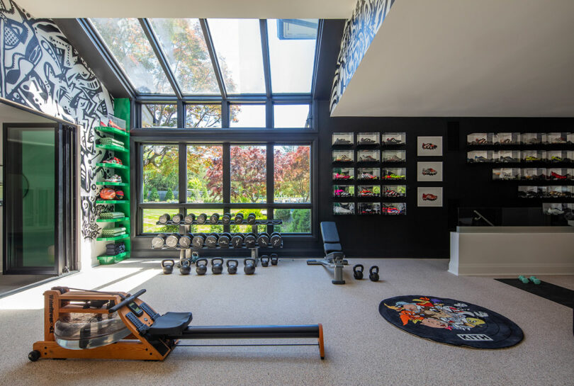 A modern home gym with a rowing machine, free weights, green shelves with sports equipment, and a wall displaying sneakers under large skylight windows.