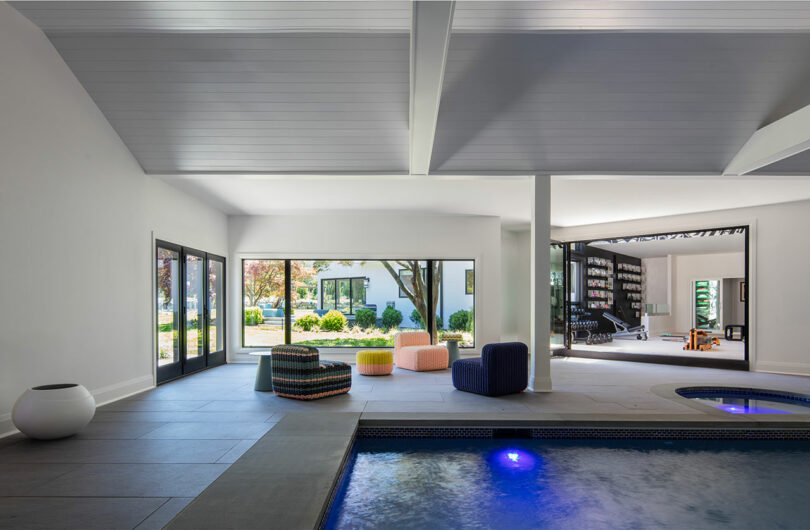 Modern indoor pool area with lounge seating, large windows, and doors opening to a garden; adjacent room visible with bookshelves and additional seating.