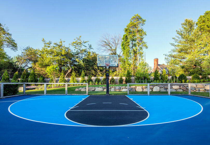 Outdoor basketball court with blue and black painted surface, surrounded by a stone wall, trees, and greenery under a clear sky.