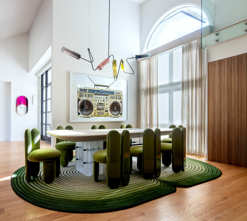 A modern dining room with a large white table, eight green chairs, a green rug, a colorful light fixture, a large boombox artwork, and floor-to-ceiling window with sheer curtains.