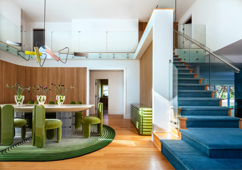Modern dining room with green chairs, oval table, decorative vases, striped rug, wooden floor, and glass staircase with blue carpet leading to an upper level.