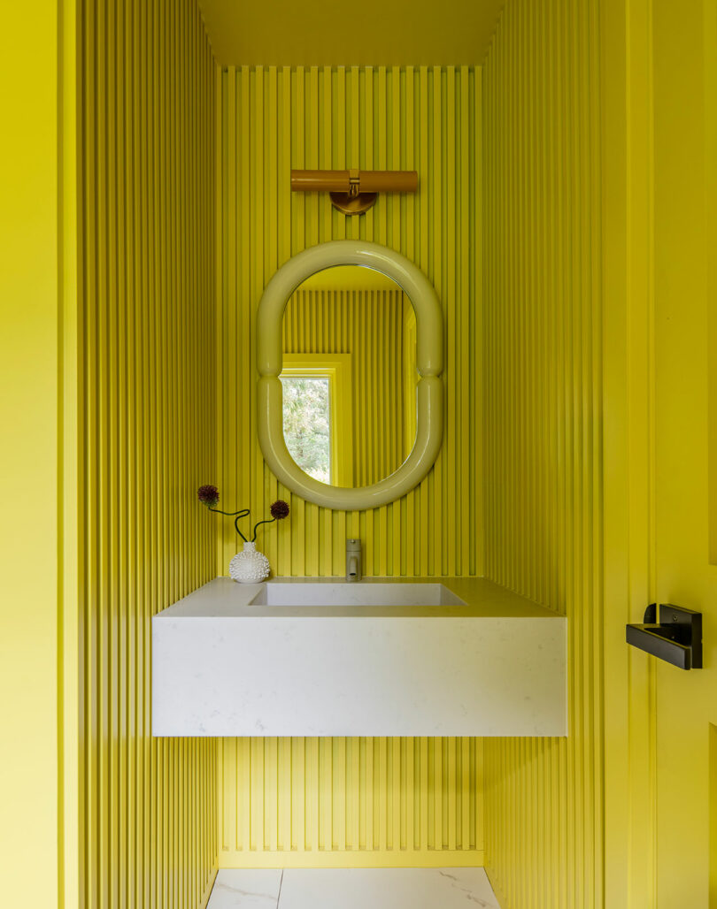 A small bathroom with bright yellow ribbed walls, a white floating sink, an oval mirror, a modern wall light, and a small vase with flowers on the counter.