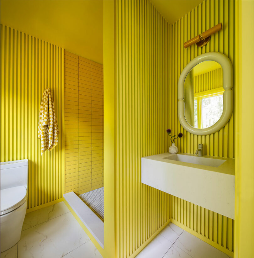 Modern bathroom with yellow vertical wall panels, a white sink with an oval mirror, a towel on the wall, and a walk-in shower with matching yellow tiles.